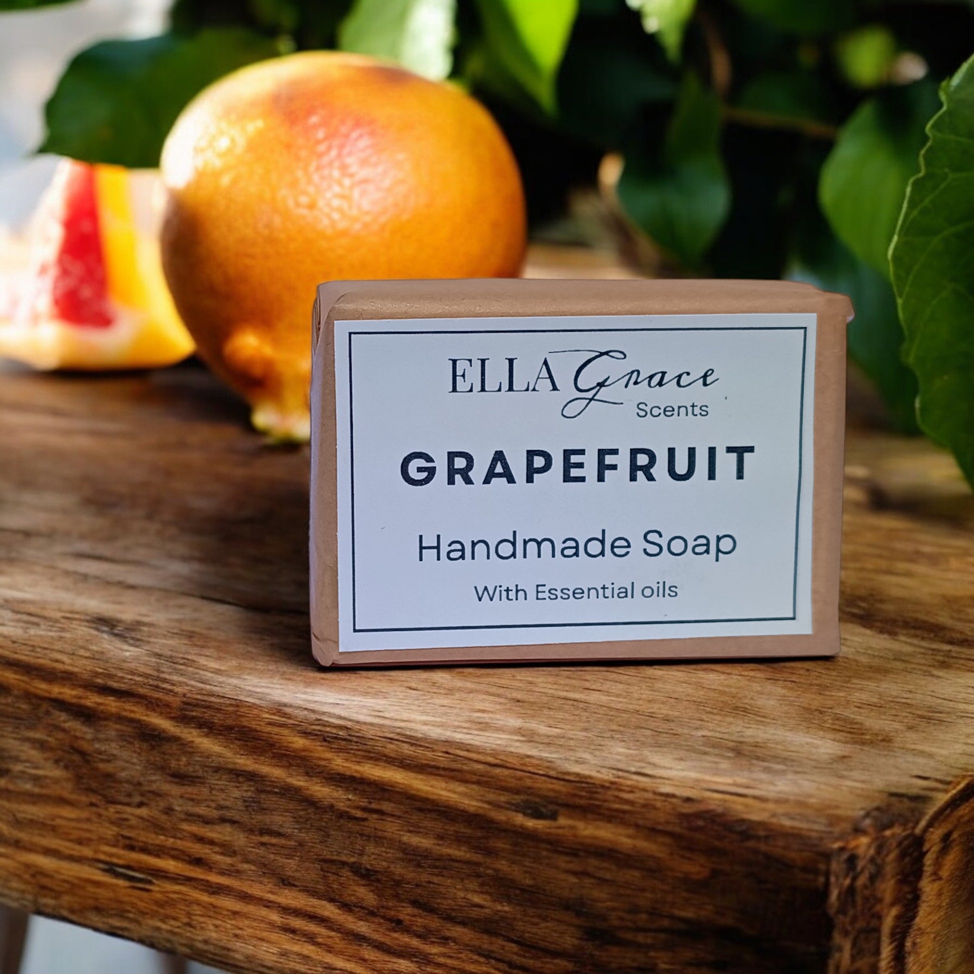 Handmade soap bar labeled 'Ella Grace Scents Grapefruit' on a wooden surface with fruits in the background.
